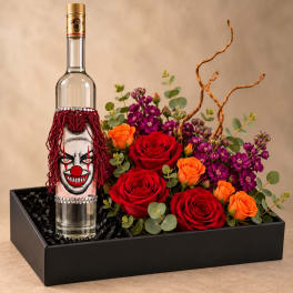 Red and orange roses with purple flowers arranged beside a decorated bottle in a black box