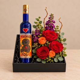 Roses and purple flowers arranged in a black gift box beside a decorated bottle