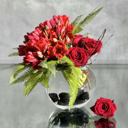Red roses and tulips arranged in a round glass vase with fern foliage.