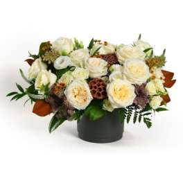 Low arrangement of cream roses with brown seed pods in a dark cylinder vase