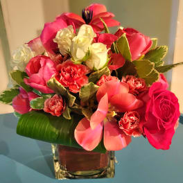 Compact cube vase arrangement of pink tulips, roses, and carnations with white roses and decorative foliage.