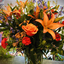 Tall mixed arrangement with orange lilies, an orange rose, red blooms and blue delphinium in a clear glass vase