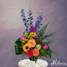 Tall mixed arrangement with blue delphinium, yellow sunflowers, orange roses, and pink blooms in a black vase.