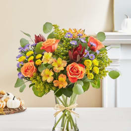 Mixed bouquet of orange roses, purple daisies, and yellow flowers in a glass vase