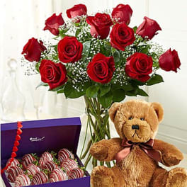 Red roses in a glass vase with a teddy bear and a box of chocolate-covered strawberries
