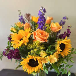 Bouquet of sunflowers, peach roses, and purple flowers in a glass vase