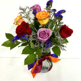 Mixed bouquet of red, yellow, and lavender roses with purple filler flowers in a glass vase with colorful ribbons