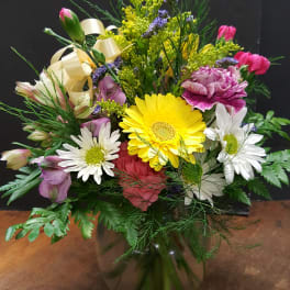 Mixed arrangement of yellow gerbera, white daisies, and pink carnations in a clear glass vase with ribbon.