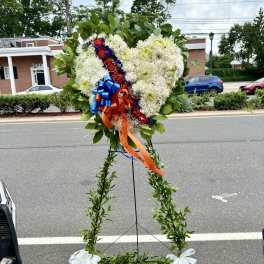 Heart-shaped floral standing spray with white flowers, red roses, and blue-orange ribbons
