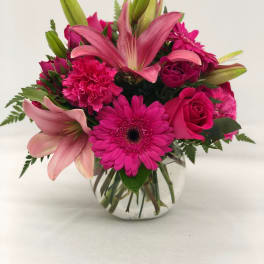 Bright pink lilies, gerbera daisies, roses, and carnations in a clear round glass vase