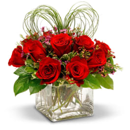 Red roses arranged in a clear square vase with looping grass accents