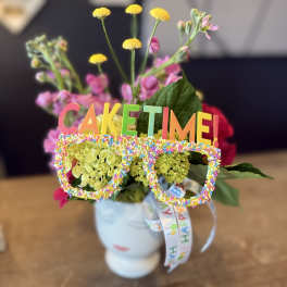 Flower arrangement in a white mug with a colorful "CAKE TIME!" topper