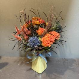 Orange roses and blue thistle in a yellow vase with a blue ribbon