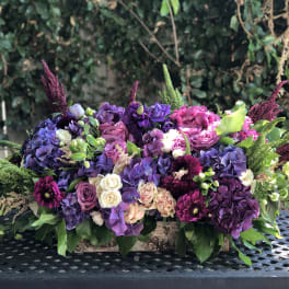 Low floral arrangement of purple, pink, and white blooms in a wooden box