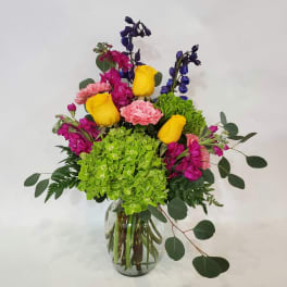 Mixed bouquet with yellow roses, pink carnations, green hydrangea, and blue flowers in a glass vase
