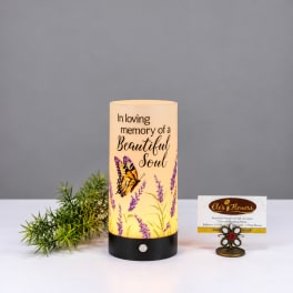Memorial candle with butterfly and lavender design beside greenery and a small card holder