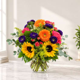 Bright mixed bouquet in a clear glass vase with sunflowers, roses, and purple blooms