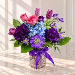 Purple and pink flower arrangement in a textured glass vase with a ribbon