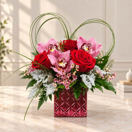 Pink orchids and red roses in a red glass vase with arched grass loops