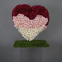 Heart-shaped rose arrangement in red, pink, and white on a grass-covered stand