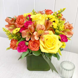 Bright arrangement of yellow roses, orange alstroemeria, and pink blooms in a green cube vase.
