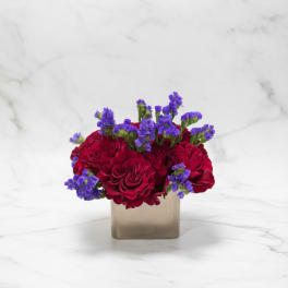 Red roses and purple filler flowers in a square gold vase