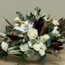 White roses and lilies in a low beige container with dried accents