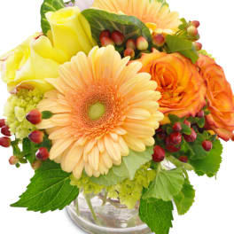 Round arrangement of peach gerbera daisy, yellow and orange roses with berries in a clear glass vase