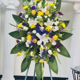 Tall standing spray of white lilies, yellow roses and purple blooms with large green leaves on an easel