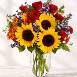 Bouquet of sunflowers, red roses, and purple daisies in a clear glass vase