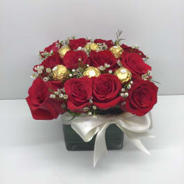 Compact arrangement of red roses and gold-wrapped chocolates in a square vase with a white bow
