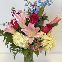 Bouquet of pink lilies, red roses, blue delphinium, and pale hydrangeas in a glass vase