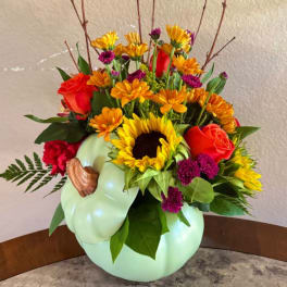 Mixed bouquet with sunflowers, roses, and orange daisies in a pale green pumpkin vase