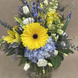 Mixed bouquet with yellow gerbera, blue hydrangea, and white blooms in a glass vase