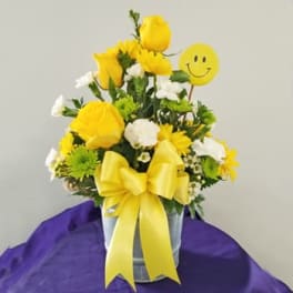 Yellow and white flower arrangement in a metal bucket with a smiley-face pick