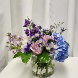Mixed bouquet in a clear glass vase with blue hydrangea, pink roses, and purple flowers