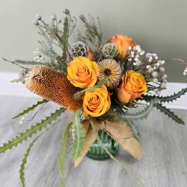 Orange roses in a glass vase with dried seed pods and a burlap ribbon