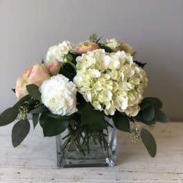 Low arrangement of white hydrangeas and pale roses in a clear glass cube vase