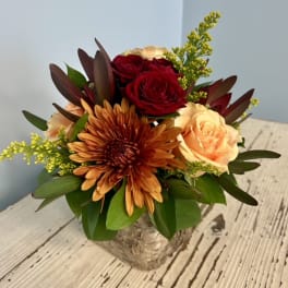 Compact bouquet of red and peach roses with an orange chrysanthemum in a birch container.