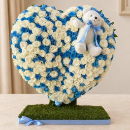 Heart-shaped rose arrangement with a teddy bear and blue ribbon on a grass-covered base