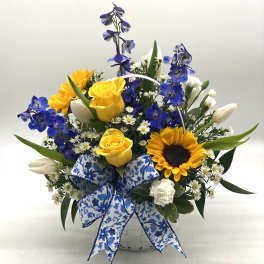 Basket arrangement with yellow roses, sunflowers, blue flowers, and white blooms