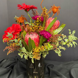 Bright mixed bouquet with red gerberas, pink protea, and orange spider accents in a dark vase