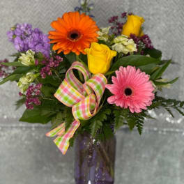 Colorful bouquet of gerbera daisies and roses in a purple vase with a plaid ribbon