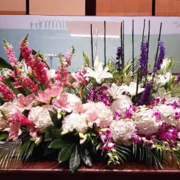 Large low centerpiece of pink lilies, white hydrangeas, and purple flowers along a stage edge