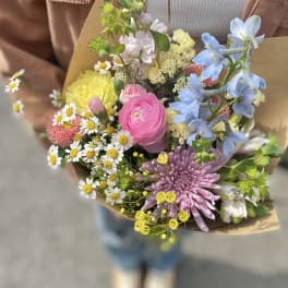 Mixed bouquet with pink, blue, yellow, and white flowers wrapped in brown paper
