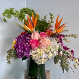Tall mixed arrangement with birds of paradise, roses, hydrangeas, and orchids in a green-lined clear glass vase.