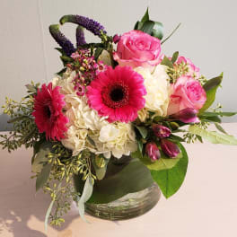 Low round arrangement of pink gerbera daisies, pink roses, and white hydrangeas in a clear glass vase