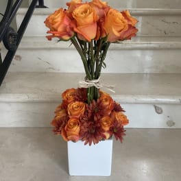 Orange roses and burgundy daisies arranged in a white square vase