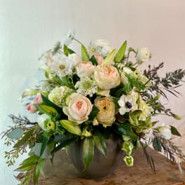 Low arrangement of peach roses and white blooms in a dark ceramic vase on a wooden surface