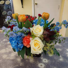 Colorful mixed bouquet with tulips, roses, hydrangeas, and blue delphinium in a low vase